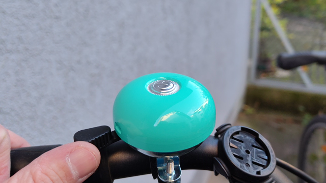 Eine leuchtend türkisfarbene Fahrradklingel wird mit dem Daumen betätigt.