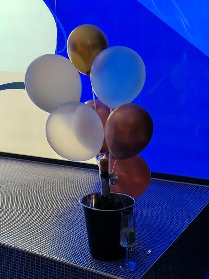 Ein Bündel Luftballons vor einem Flaschenkühler mit einer Flasche Sekt. 