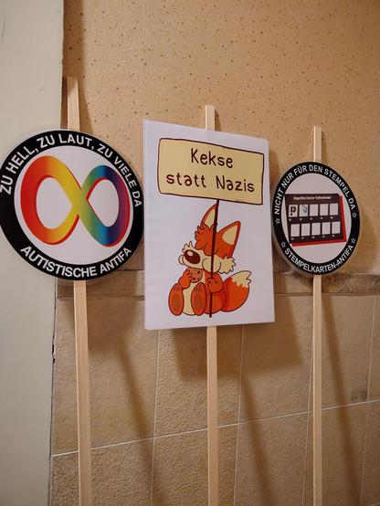 Drei auf Holzstäbe montierte Plakate lehnen an einer Wand. 
Eines zeigt einen Comic-Fuchs und die Aufschrift "Kekse statt Nazis". Eines hat das Autismus-Regenbogen-Unendlichkeitssymbol im Zentrum und die Aufschrift "Zu hell, zu laut, zu viele da - Autistische Antifa. 
Eines zeigt eine Prüf-Stempelkarte im Zentrum und hat die Aufschrift "nicht nur für den Stempel da - Stempelkarten- Antifa"