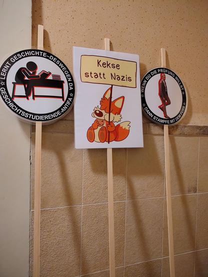 Drei auf Holzstäbe montierte Plakate lehnen an einer Wand. 
Eines zeigt einen Comic-Fuchs und die Aufschrift "Kekse statt Nazis".
Eines hat das Icon einer am Tisch lesenden Person im Zentrum und die Aufschrift "Lernt Geschichte-deswegen da Geschichtsstudiende-Antifa" 
Eines hat das Bild einer aufstampfenden Alice Weidel im Zentrum und die Aufschrift "Wenn du die Prüfung willst dann stampfe mit dem Fuß". 