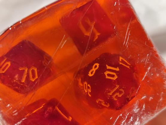 Ein rötlich-durchsichtiges Seifenstück. Darin eingebettet sieht man mehrere violette Würfel mit unterschiedlichen Flächenzahlen, wie man sie von D&D kennt. Das Seifenstück erinnert an das D&D-Monster "Gelatinous Cube". 